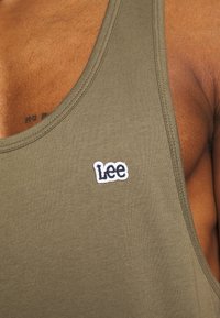 Gros plan d'une personne portant un débardeur vert olive avec un petit logo "Lee" blanc et noir sur la poitrine.