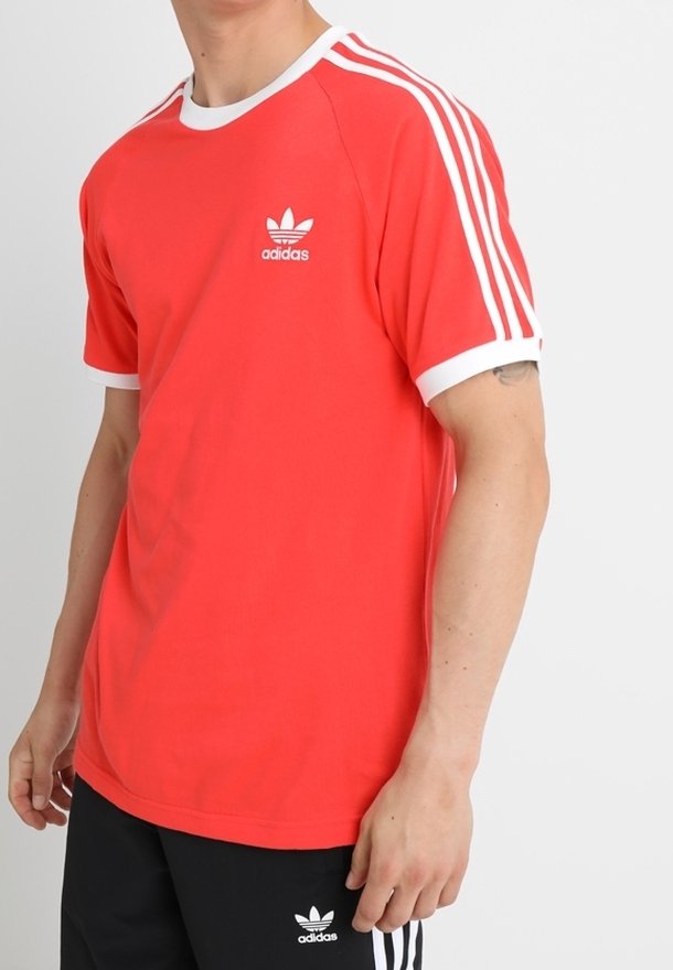 Punainen lyhythihainen t-paita, jossa on valkoinen reunus ja kolme valkoista raitaa kummallakin olkapäällä. Rintaan on painettu pieni valkoinen Adidas-logo.