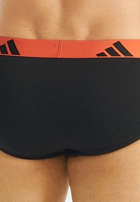 Mustat uimapuvut punaisella joustavalla vyötäröllä, jossa on mustia logoelementtejä. Kangas näyttää sileältä ja istuu hyvin.