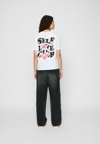 Tricou alb din bumbac cu text negru îndrăzneț "SELF LOVE CLUB" și accente florale roz pe spate. Asortat cu pantaloni supradimensionați de culoare gri închis.