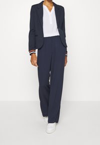 Blazer bleu marine avec revers et poignets rayés, associé à un pantalon large bleu marine et des baskets blanches. Comprend une chemise blanche avec un col en V.