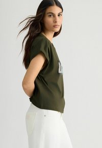 T-shirt verde oliva a maniche corte con una vestibilità rilassata e una grafica decorata con cristalli sul davanti, abbinato a pantaloni bianchi.