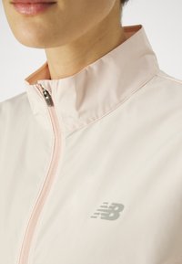 Giacca zip-up rosa chiaro con colletto alto e finitura testurizzata. Presenta un logo New Balance grigio discreto sul petto.