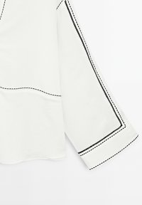 Blouse blanche avec des coutures contrastées noires le long des coutures et des poignets. Les longues manches évasées confèrent une forme distinctive au design.