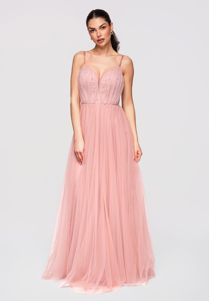 Femme en robe sans manches en tulle rose avec corsage perlé et décolleté plongeant en V, debout devant un fond clair uni.