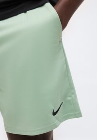 Άτομο που φοράει ανοιχτοπράσινα σορτς Nike με μαύρο λογότυπο Swoosh στο δεξί πόδι, χέρι στην τσέπη σε λευκό φόντο.