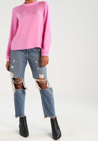Maglia rosa a maniche lunghe con scollatura rotonda, abbinata a jeans blu strappati con ampie aree strappate e bordi sfilacciati. Stivaletti neri alla caviglia.