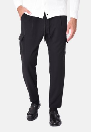 Pantaloni cargo neri con tasche laterali, vita regolabile con cordoncino e design a gamba affusolata. Indossati con scarpe nere e un cardigan bianco.