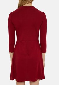 Czerwona sukienka do kolan wykonana z miękkiego materiału, z rękawami o długości trzech czwartych, okrągłym dekoltem oraz dopasowaną górą i rozkloszowanym dołem.