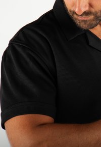 T-shirt polo noir en tissu texturé, avec un col et des manches courtes. La coupe est ajustée au niveau des épaules.