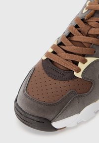 Gray and brown sneaker with a padded design, featuring perforated accents, flat laces, and a rubber sole. Textured leather and suede materials.