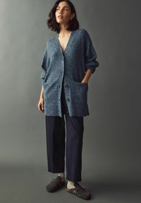 Blå stickad cardigan med långa ärmar, två framfickor och knappar. Kombinerad med mörka byxor och bruna träskor. Neutral bakgrund.
