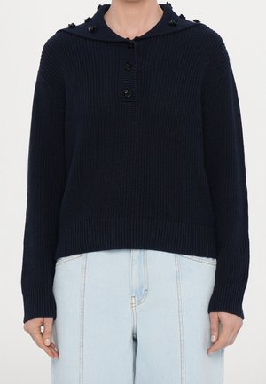 Jersey de punto azul marino con botones negros en forma de flor en el cuello y vaqueros de cintura alta y color azul claro con costuras visibles.