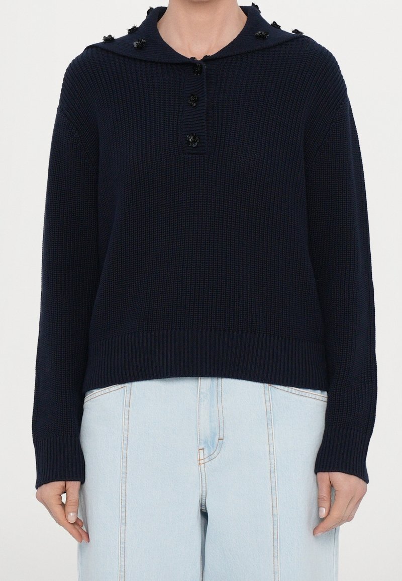 Pull en tricot bleu marine avec des boutons noirs en forme de fleurs sur le col et jean taille haute bleu clair avec coutures visibles.