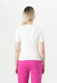 Žena s dlhými blond vlasmi, oblečená v jednoduchom bielom tričku s krátkym rukávom a jasnoružových nohaviciach, videná zozadu na bielom pozadí.