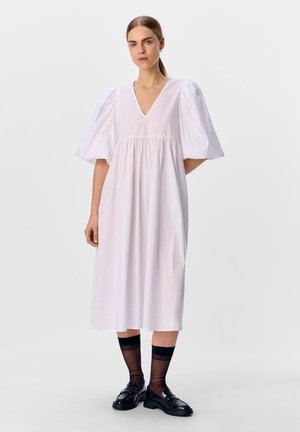 Weißes Midi-Kleid mit V-Ausschnitt, Puffärmeln, gerafftem Taillenschnitt und fließender Silhouette. Getragen mit schwarzen Loafers und transparenten Socken.