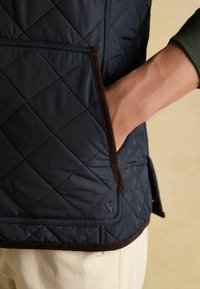 Námornícky prešívaný vest s hnedými kordovým lemovaním a bočnou kapsou, s jemným detailom loga. Textúrovaný, polstrovaný materiál.