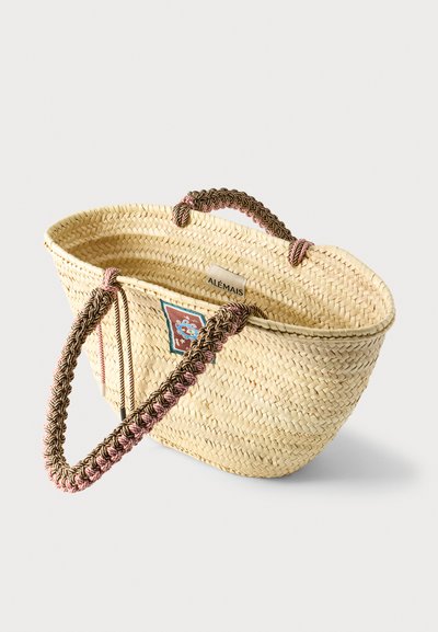 Πλεκτή τσάντα από ψάθα με πλεγμένες καφέ και ροζ χειρολαβές και ένα μικρό πολύχρωμο σήμα στην εξωτερική πλευρά.