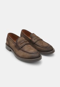 Mocasines de ante marrón con una textura suave, que presentan una puntera redondeada cosida y una tira decorativa a través del empeine. Suela de goma.