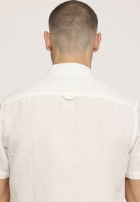 Chemise en lin blanc, à manches courtes, détails de couture visibles aux épaules, texture subtile, boucle au niveau du cou. Vue de dos.