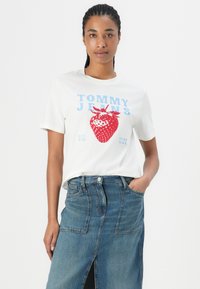 T-shirt en coton blanc avec un graphique de fraise rouge et le texte "TOMMY JEANS" en bleu. Associé à une jupe en denim avec deux poches avant.