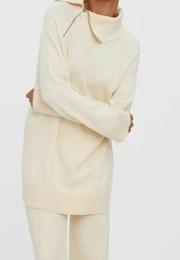 Personne portant un pull oversize couleur crème avec un col montant et un détail zippé, assorti à un pantalon crème.