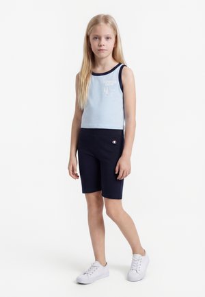 Ξανθιά κοπέλα που στέκεται, φοράει ανοιχτό μπλε αμάνικο τοπ με το κείμενο "Summer Camp" και σκούρα μπλε σορτς με λογότυπο, λευκά παπούτσια.
