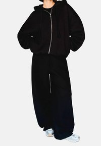 Czarna, oversize, zapinana na zamek bluza z kapturem z trokami, w połączeniu z luźnymi czarnymi spodniami dresowymi. Materiał wydaje się miękki, odpowiedni do codziennych stylizacji.