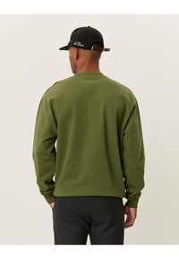 Felpa verde oliva con scollatura rotonda e maniche lunghe. Tessuto morbido, design minimalista e vestibilità comoda. Cappellino nero indossato.