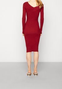 Robe en tricot rouge à manches longues, silhouette ajustée, dos en V, et texture côtelée ; associée à des sandales blanches à bout ouvert.