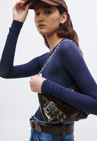 Bolso bandolera de charol marrón con tachuelas plateadas y detalle de hebilla, llevado sobre una camiseta ajustada de manga larga azul marino y vaqueros azules.