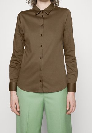 Camicione marrone a maniche lunghe con colletto, apertura frontale con bottoni e texture liscia; abbinato a pantaloni leggeri verdi.