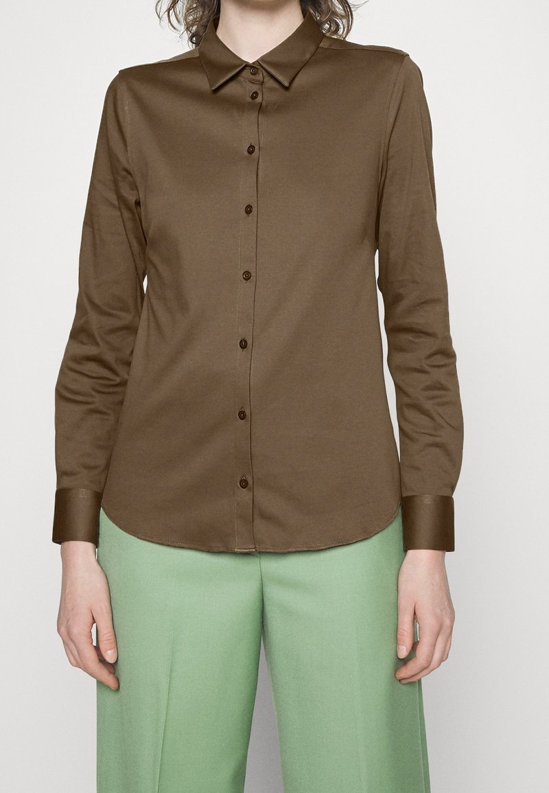 Camicione marrone a maniche lunghe con colletto, apertura frontale con bottoni e texture liscia; abbinato a pantaloni leggeri verdi.