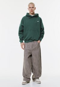 Grüne Kapuzenjacke mit einer Fronttasche und kleinem Logo, kombiniert mit übergroßen grauen gestreiften Hosen und hellen Sneakers.