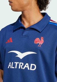 Blå sports polo skjorte med røde accenter, prydet med et kyllingemblem og "ALTRAD" logoen i hvid. Kravedesign tilføjer tekstur.
