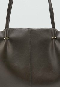Bolso tote de cuero marrón oscuro con una textura suave, que presenta un diseño minimalista, ligeras pliegues y acentos en dorado.