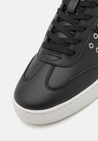 Sneaker in pelle nera con cuciture tono su tono, lacci neri rotondi e occhielli metallici. La suola è bianca con una superficie testurizzata.