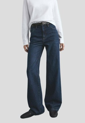 Jeans de mezclilla oscuros de pierna ancha y cintura alta, con bolsillos frontales y sutiles detalles de costura, combinados con una camisa blanca de mangas largas.