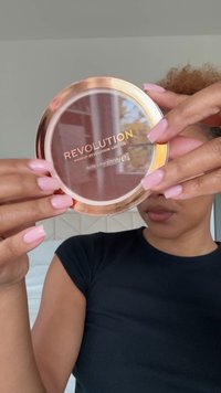 Rund makeup-kompakt med klart lokk, gullkant og preget "REVOLUTION"-tekst. Inneholder bronsepudder, vist mot en nøytral bakgrunn.