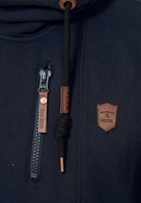 Mornarska dukserica s patentnim zatvaračem i pletenicom za podešavanje. Sadrži smeđe kožne akcentne detalje i oznaku s nazivom brenda "INDICODE." Mekana tekstura.