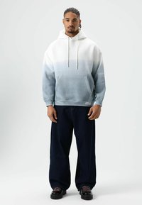 Hoodie met een verloop van wit naar lichtblauw, de stof is zacht, heeft een voorzak, gecombineerd met donkerdere wijdere jeans en zwarte schoenen.