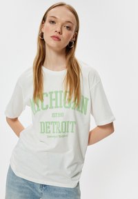 Hvid bomulds t-shirt med grøn tekst: "MICHIGAN EST 1990 DETROIT University of California." Korte ærmer og afslappet pasform.