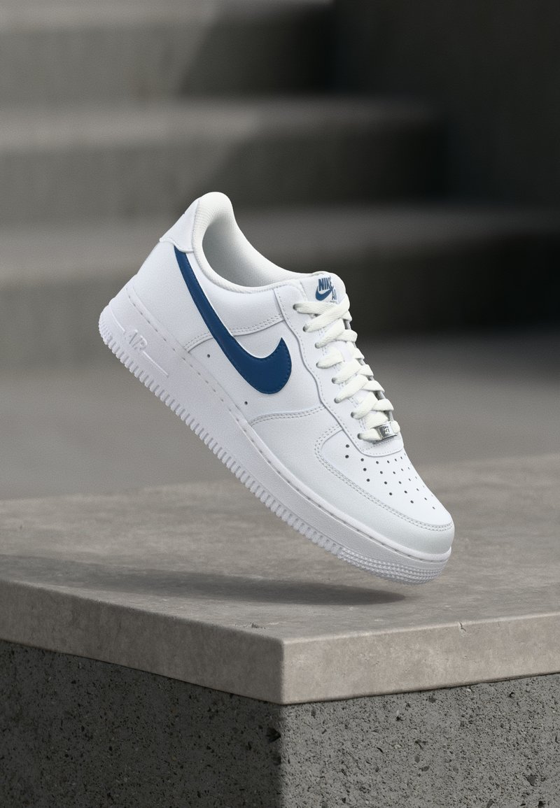 Λευκό αθλητικό παπούτσι Nike Air Force 1 με μπλε λογότυπο swoosh που αιωρείται πάνω από μια γκρι τσιμεντένια άκρη.