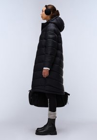 Zwarte pufferjas met capuchon, met een horizontaal gestikt ontwerp. Model draagt zwarte leggings en militaire laarzen met veters. Oorkappen zijn inbegrepen.