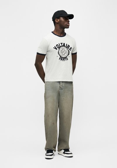 Άνδρας που φοράει μαύρο καπέλο, λευκό μπλουζάκι με γραφικά, φαρδιά ξεβαμμένα τζιν και ασπρόμαυρα αθλητικά παπούτσια, στέκεται με τα χέρια πίσω από την πλάτη.