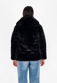 Giacca in finta pelliccia nera con colletto alto, caratterizzata da una texture morbida e una vestibilità ampia. Indossata con jeans in denim blu chiaro.