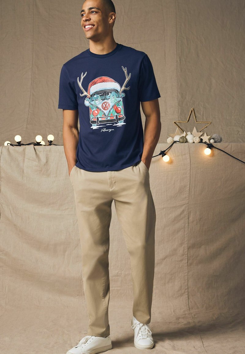 Marineblå t-shirt med et farverigt grafik af en VW-bus med gevir og en julestråhat, parret med lyse khaki-bukser og hvide sneakers.