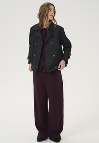 Cappotto trench nero doppiopetto con grandi bottoni, spalline, indossato su una blusa bordeaux e pantaloni larghi. Tessuto morbido, vestibilità su misura.