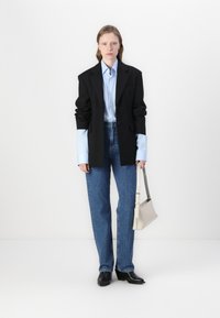 Črn blazer, svetlo modra srajca z dolgimi rokavi, široke modre kavbojke, črne čevlje, drži majhno bež torbico, minimalistična podlaga.