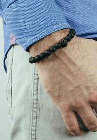Bracciale in pietra lavica nera con perle tonde, indossato su un polso, con una camicia blu a fantasia visibile sullo sfondo.